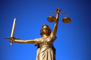 Eine bronzene Justizia-Statue vor blauem Hintergrund. Justizia hält in der einen Hand eine Waage, in der anderen ein Schwert.
